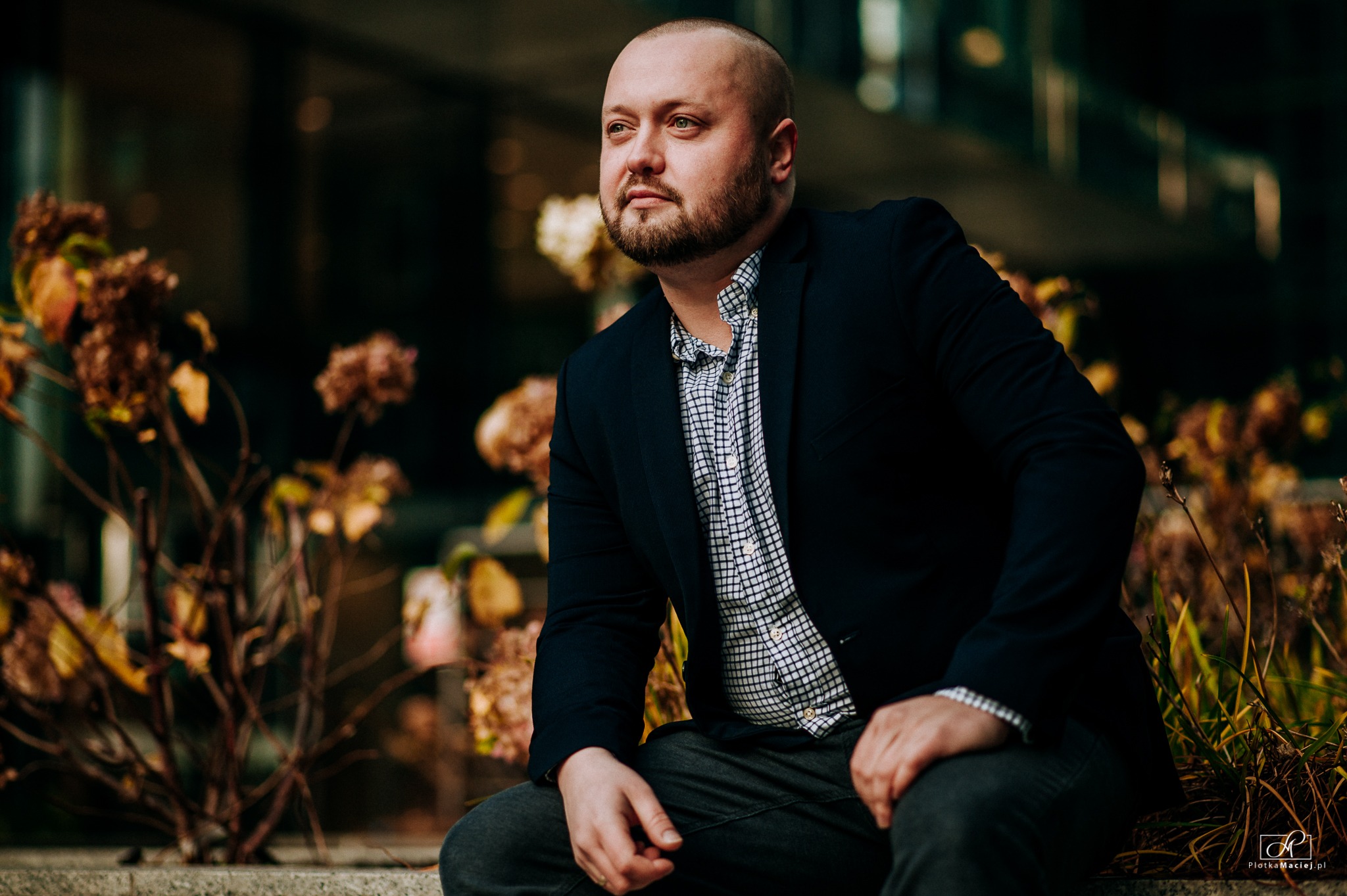 Portret mężczyzny w marynarce i kraciastej koszuli, siedzącego na tle nowoczesnego budynku i uschniętych kwiatów, spoglądającego w lewo.