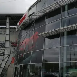 Elewacja salonu samochodowego Toyota z dużymi szklanymi oknami i plakatem czerwonego samochodu, widoczna wieża telekomunikacyjna w tle.