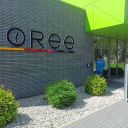 BlueService Sp. z o.o. - Pracownik w niebieskiej koszulce myje przeszklone wejście do budynku z szarą elewacją za pomocą długiego kija z systemem czyszczącym na kółkach obok, widoczny napis KOREE na elewacji.