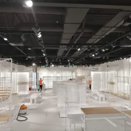 Wnętrze sklepu w trakcie porządkowania, widoczny pracownik w pomarańczowej kamizelce i odkurzacz przemysłowy na błyszczącej podłodze, oświetlenie szynowe na suficie.