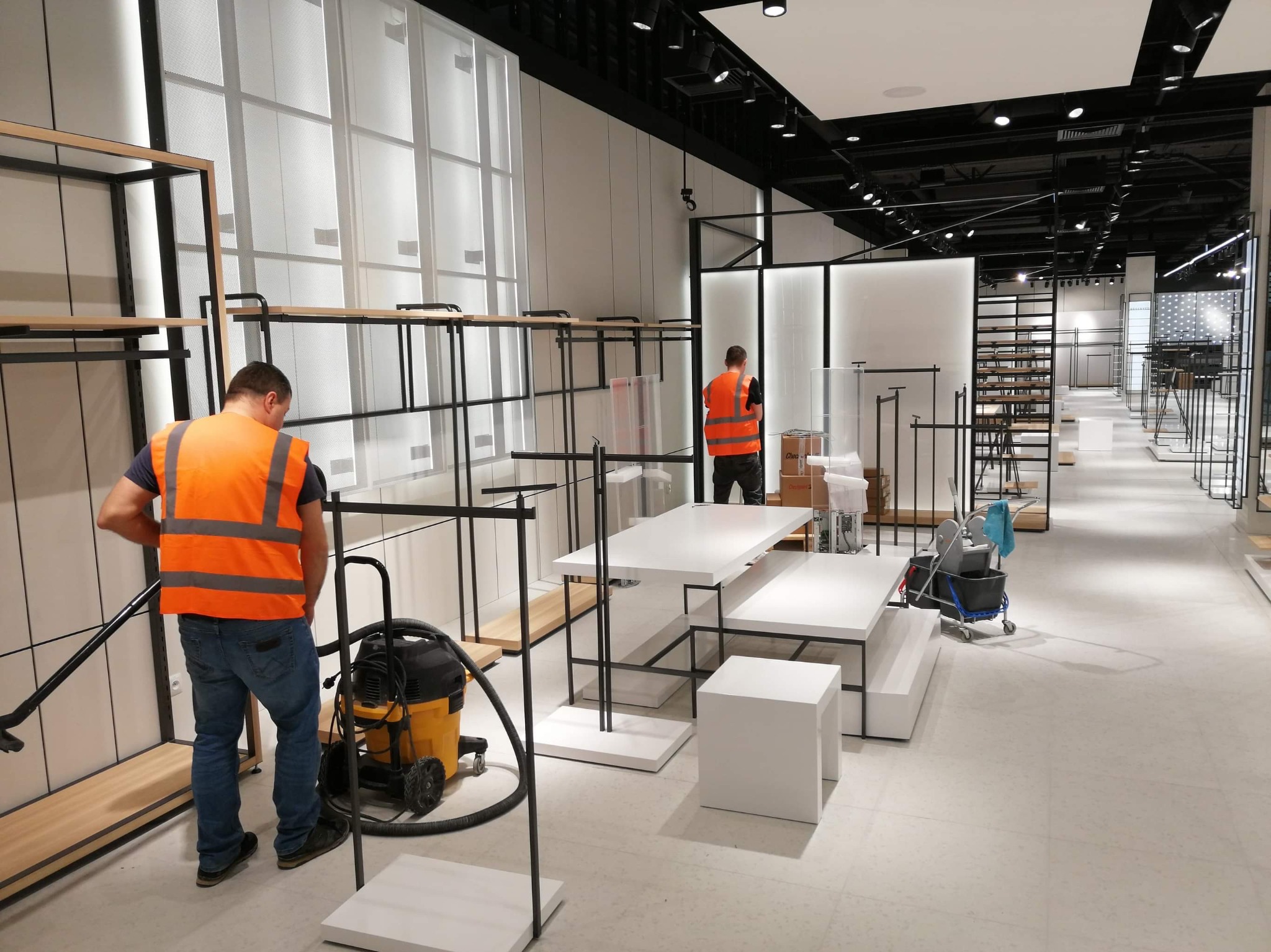 Pracownik w pomarańczowej kamizelce sprząta odkurzaczem podłogę w nowym sklepie, gdzie ustawiane są metalowe regały z drewnianymi półkami i białe stoliki.