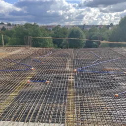 Zbrojenie stropu z widocznymi, niebieskimi rurkami peszel i pomarańczowymi puszkami instalacyjnymi, przygotowane do zalania betonem, w tle zabudowa miejska i pochmurne niebo.