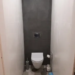 Wnętrze wykończonej łazienki z toaletą, szarą mozaiką na podłodze i ciemnoszarą ścianą z płytek za toaletą, widoczny dozownik do mydła i szczotka do WC.