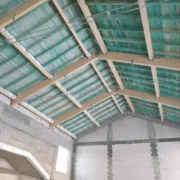 Surowy stan poddasza z widoczną konstrukcją dachu z drewnianymi belkami i deskami w kolorze zielonym, widoczne stalowe elementy mocujące, ściany z betonu komórkowego w trakcie wznoszenia.