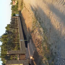 Ozdobne, czarne ogrodzenie z kamienną podmurówką i furtką, oddzielające posesję od polnej drogi. Widoczne ślady opon na ziemi i zieleń w tle. Ujęcie z dołu.