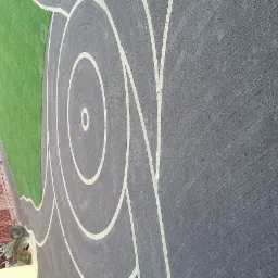 Efektowna nawierzchnia z kostki brukowej ułożonej w spiralny wzór, sąsiadująca z zielonym trawnikiem i elementami architektury ogrodowej. Widok z góry.