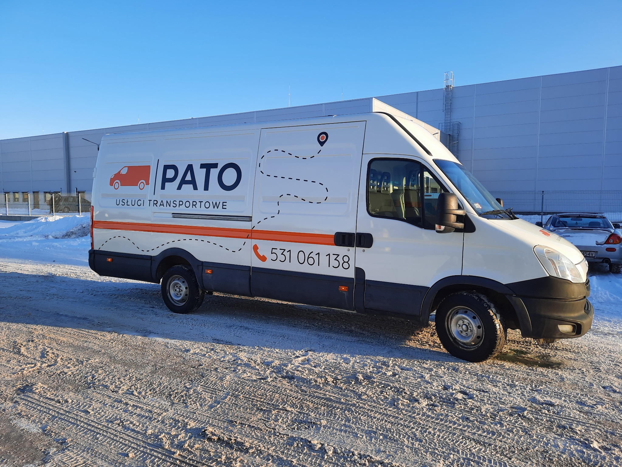 Biały bus z logo firmy transportowej 'PATO' na tle magazynu i zaśnieżonego placu.