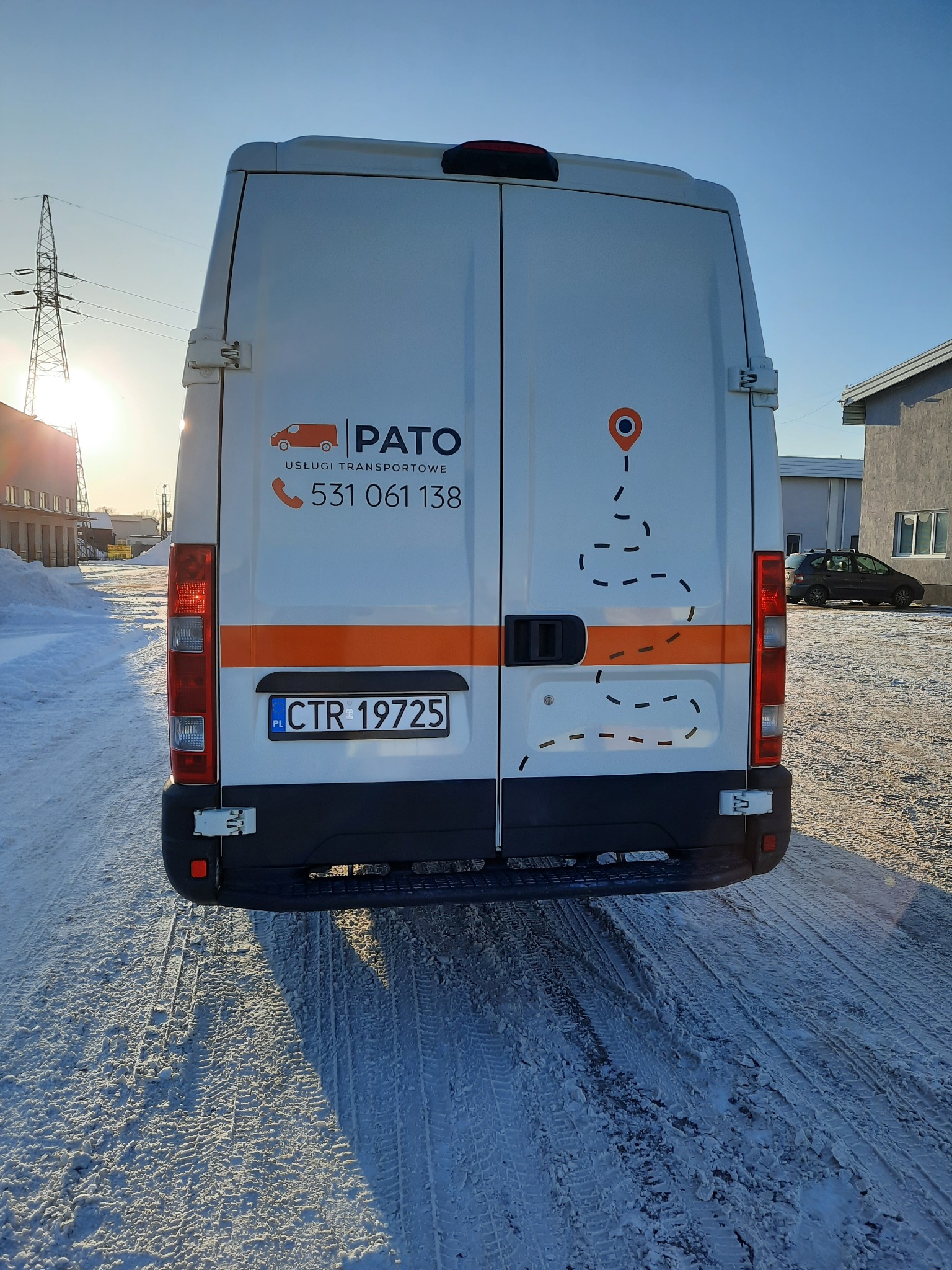 Tył białego busa transportowego z logo firmy PATO i numerem telefonu, zaparkowanego na śniegu w słoneczny dzień.