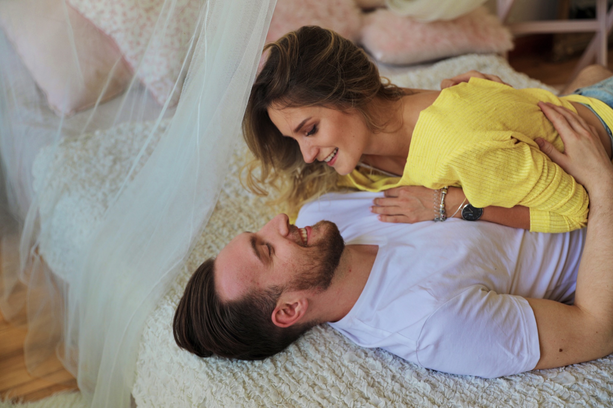 Uśmiechnięta para leżąca na łóżku pod baldachimem, kobieta w żółtej bluzce opiera się na mężczyźnie w białej koszulce.