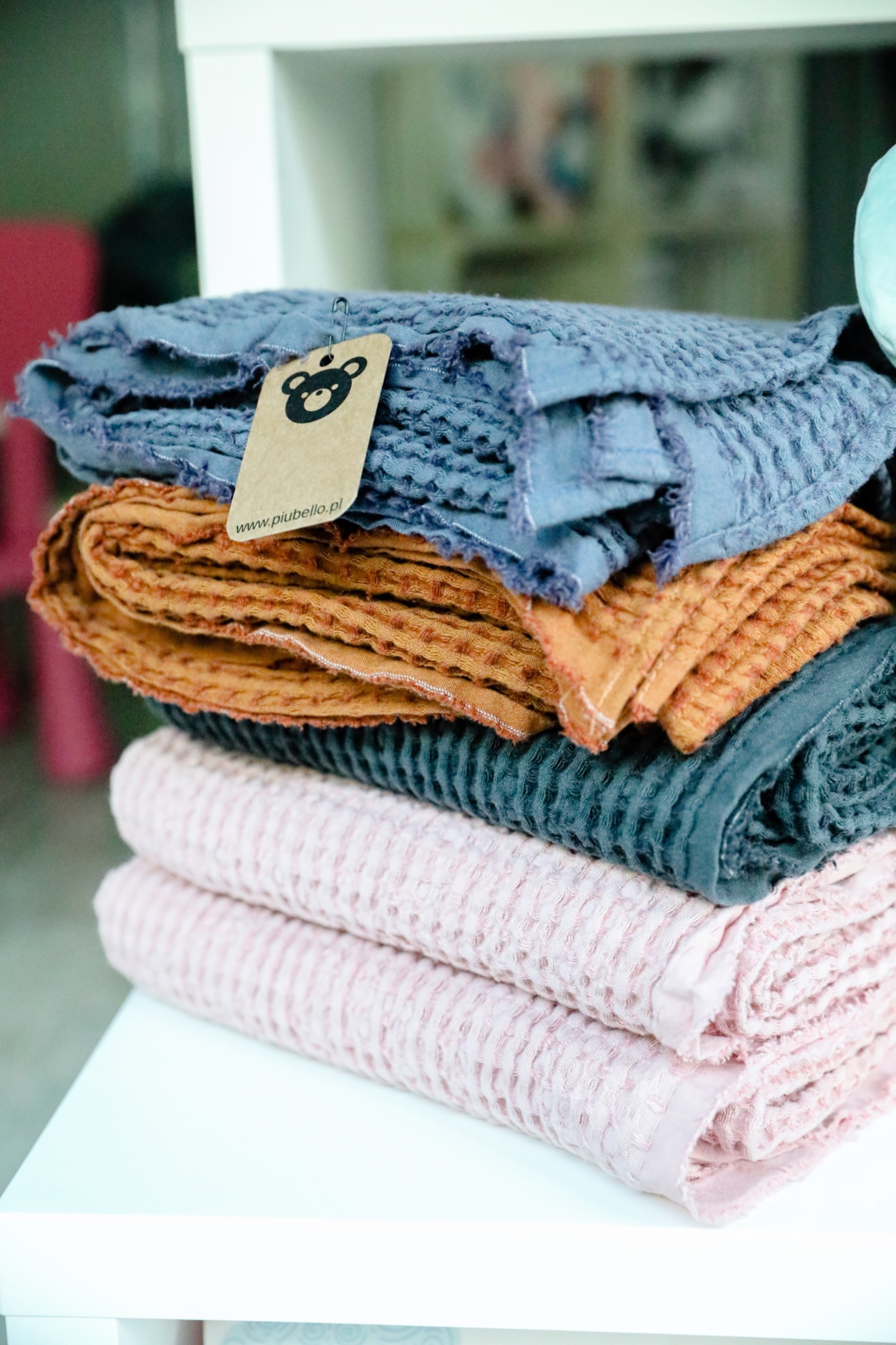 Stos bawełnianych ręczników w odcieniach różu, zieleni, pomarańczy i błękitu, z fakturą wafelkową, na białym stoliku, z etykietą marki przypiętą agrafką do górnego ręcznika.