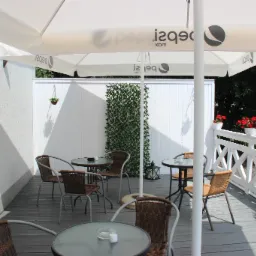 Zewnętrzny taras kawiarni z wiklinowymi krzesłami i okrągłymi stolikami pod parasolem z logo napoju, dekorowany kwiatami w doniczkach i zieloną ścianką.