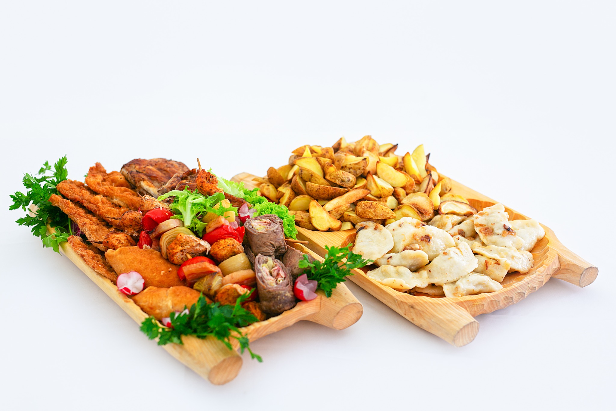 Dwie drewniane tace z różnorodnymi potrawami: kotlety schabowe, szaszłyki, roladki mięsne, pieczone ziemniaki i pierogi z okrasą, udekorowane świeżą zieloną pietruszką i rzodkiewką na białym tle.