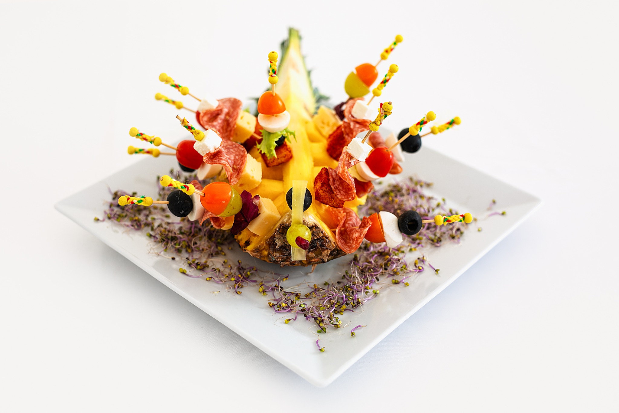 Dekoracyjna kompozycja z przekrojonego ananasa, na którym umieszczono koreczki z salami, serem, pomidorkami i oliwkami, całość na białym talerzu z kiełkami.