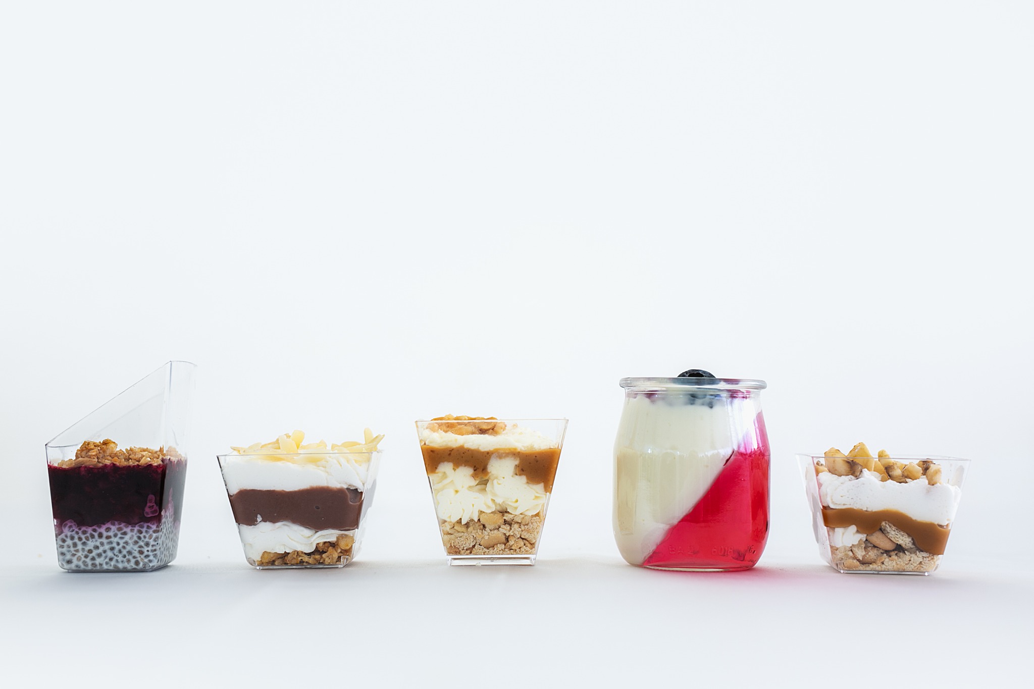 Rząd pięciu deserków w przezroczystych naczyniach o różnych kształtach, na białym tle, z warstwami musu, galaretki, owoców i posypki.