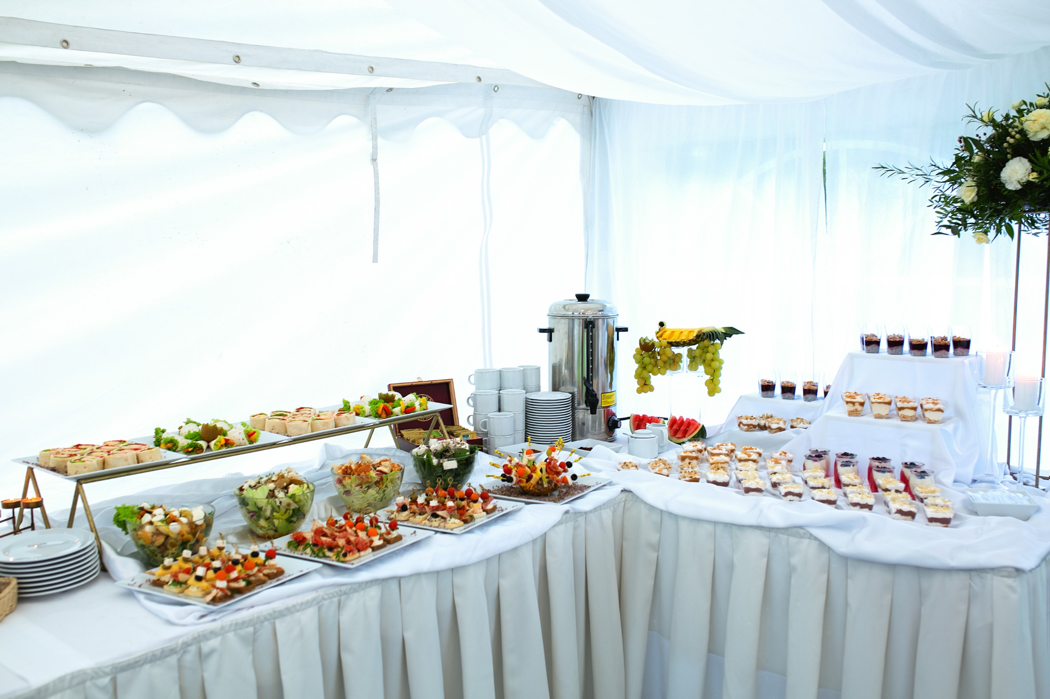 Elegancko zastawiony stół z różnorodnymi przekąskami, deserami i napojami w białym namiocie, dekoracja kwiatowa w prawym górnym rogu.