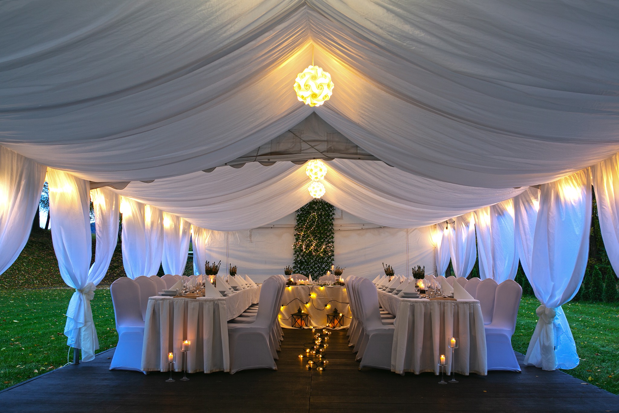 Uroczysta aranżacja stołów w białym namiocie z dekoracyjnym oświetleniem: lampiony, świece i girlanda świetlna, zielona ściana z napisem LOVE w tle.
