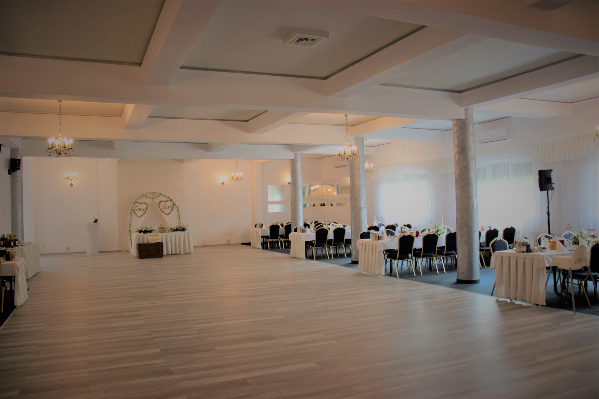 Sala bankietowa przygotowana na uroczystość, z dekoracjami kwiatowymi, nakrytymi stołami i parkietem w odcieniach szarości.