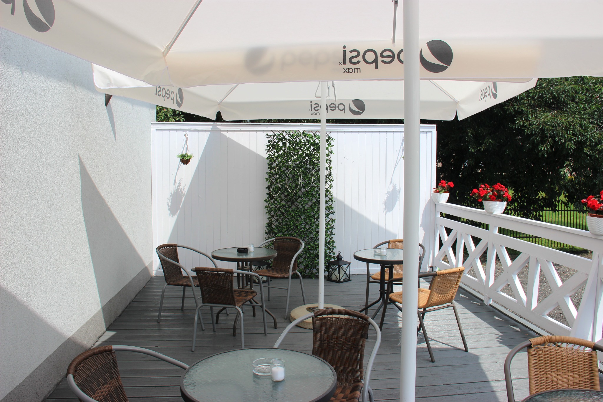 Zewnętrzny taras kawiarni z wiklinowymi krzesłami i okrągłymi stolikami pod parasolem z logo napoju, dekorowany kwiatami w doniczkach i zieloną ścianką.