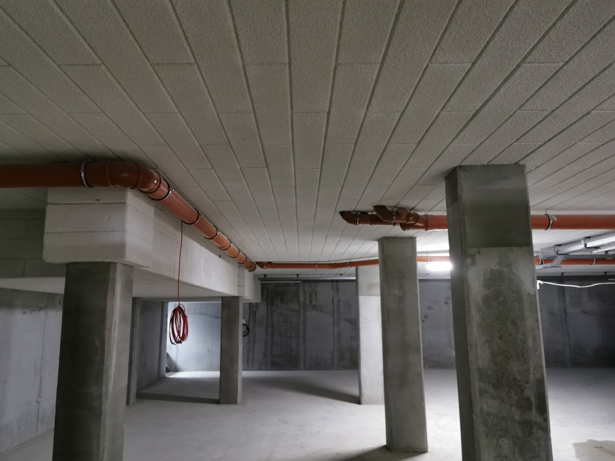 Poddasze z betonowymi kolumnami i izolacją stropu z widocznymi rurami instalacji sanitarnej.