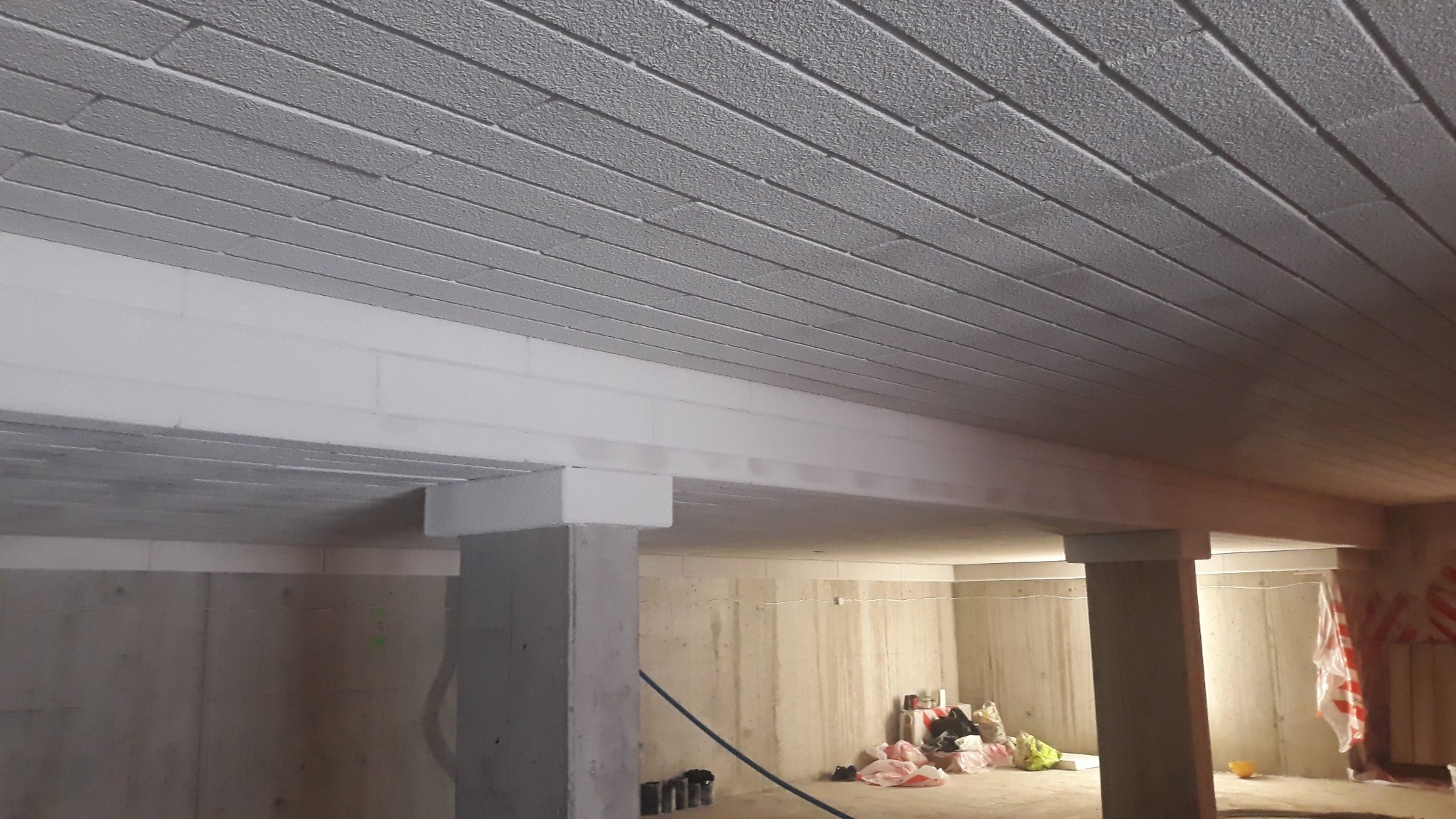 Szare, ocieplone poddasze z widocznymi belkami konstrukcyjnymi i betonowymi kolumnami w surowym stanie.