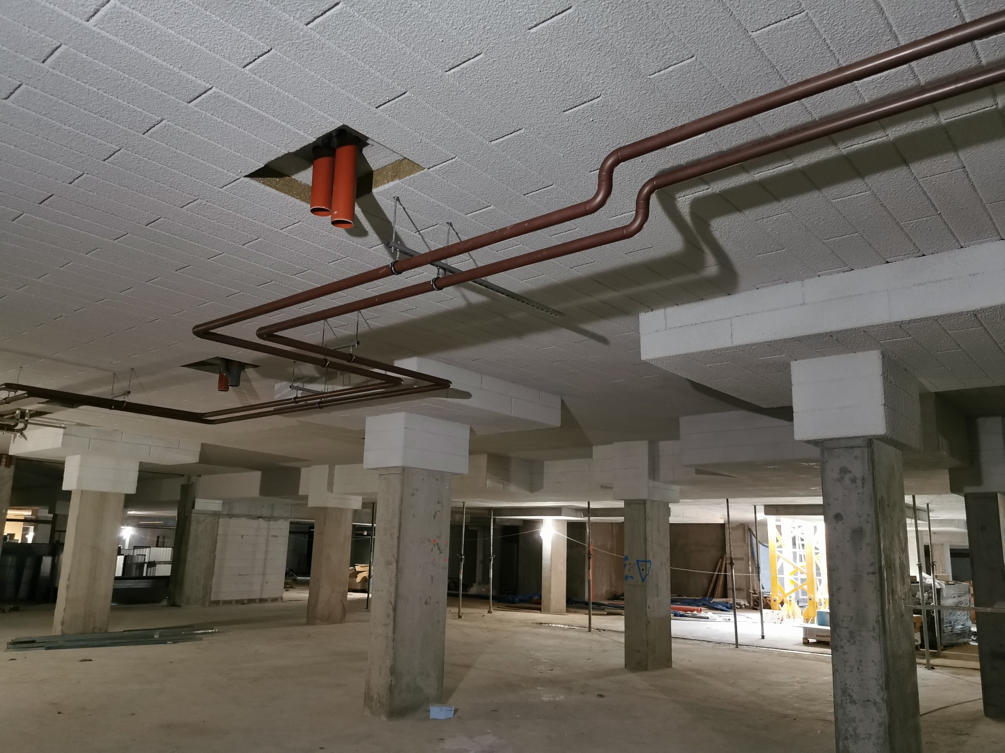 Instalacja rur wentylacyjnych i grzewczych pod stropem budynku w trakcie budowy, widoczne betonowe kolumny i pustakowy strop z widoczną fakturą.