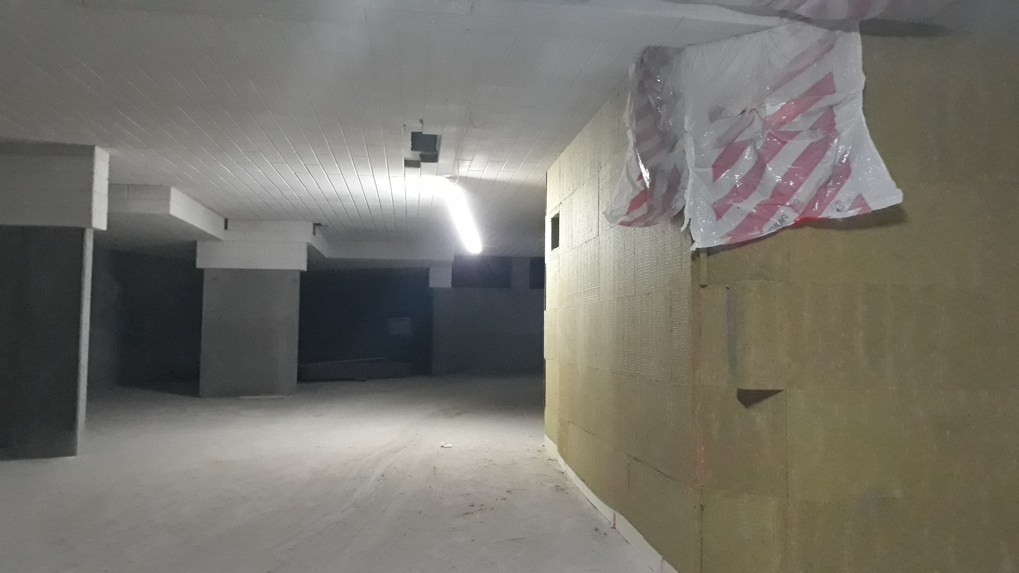 Ściana pokryta płytami izolacyjnymi, zabezpieczona folią w biało-czerwone pasy, widok na surowy betonowy strop i słupy w tle.
