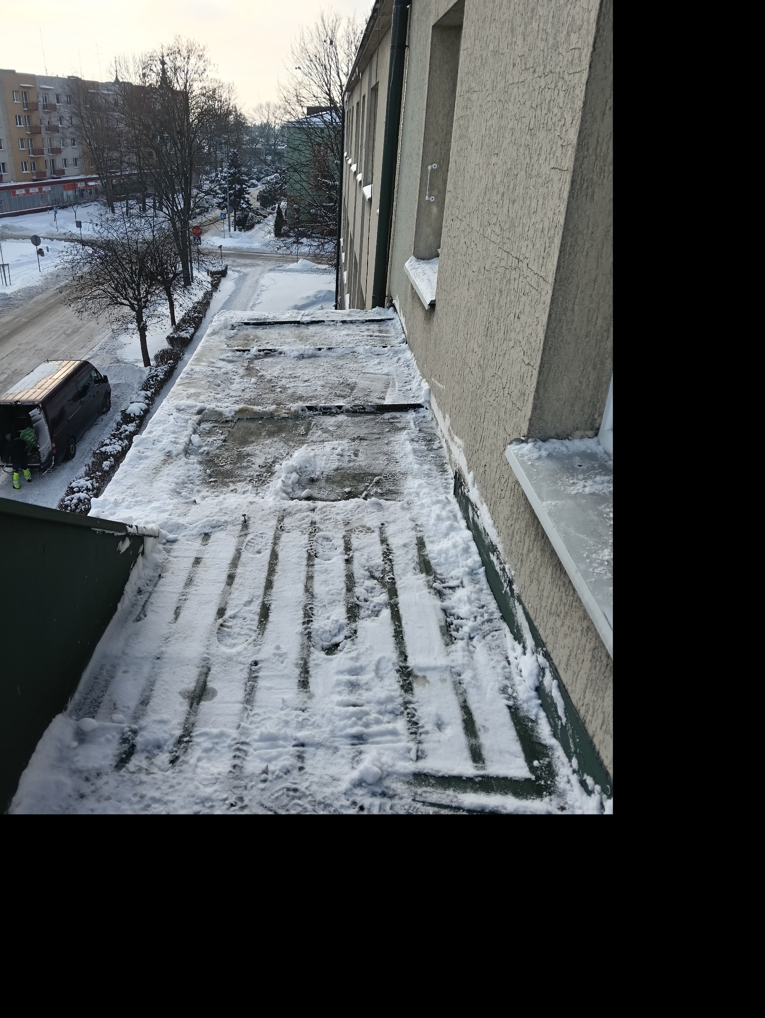 Śnieg na dachu budynku częściowo odśnieżony, widoczne ślady butów. W tle ulica i budynki mieszkalne, pracownik przy samochodzie. Perspektywa z góry.