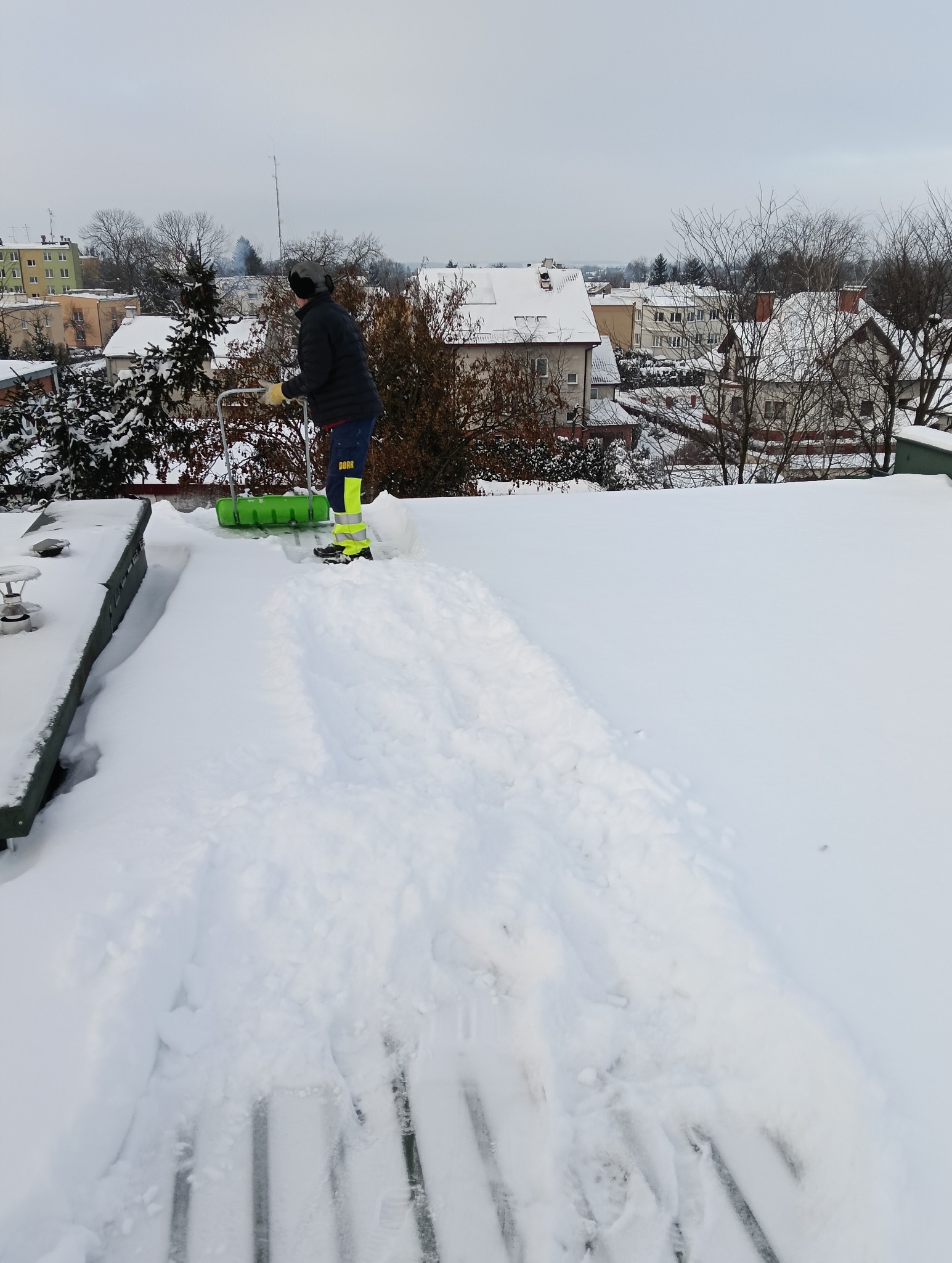 Pracownik w odzieży roboczej odśnieża płaski dach zieloną łopatą. W tle zabudowa miejska i pochmurne niebo. Widoczny fragment odśnieżonej powierzchni.