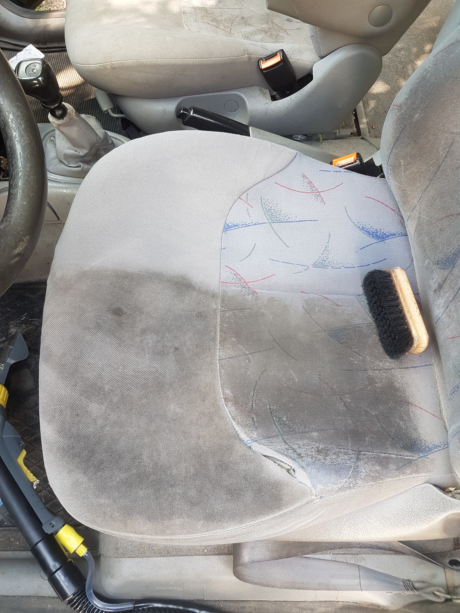 Czyszczone jasnoszare siedzenie samochodowe z widoczną różnicą w czystości po lewej stronie, obok leży szczotka i dysza odkurzacza piorącego.