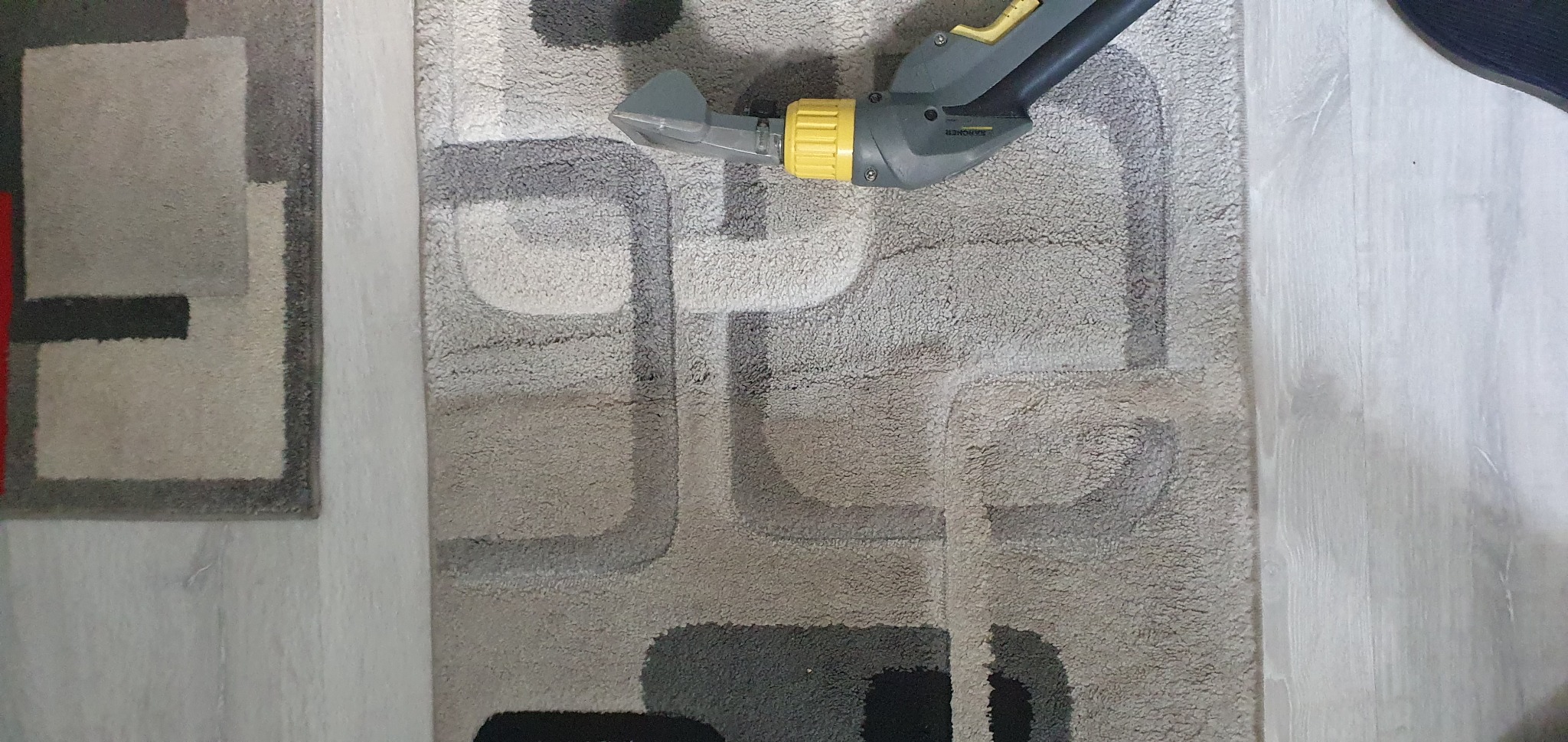 Szaro-beżowy dywan z geometrycznym wzorem czyszczony parowym odkurzaczem Karcher; widoczna dysza i fragment uchwytu urządzenia.