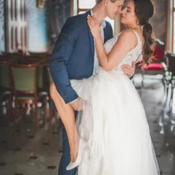 Uśmiechnięta panna młoda w koronkowej sukni, unoszona przez pana młodego w granatowym garniturze, na tle bogato zdobionego wnętrza z parkietem i dekoracyjnymi ścianami.