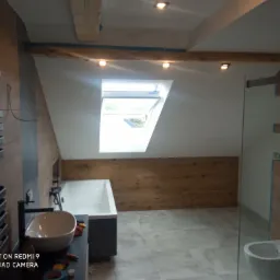 Nowoczesna łazienka na poddaszu z oknem dachowym, drewnianym wykończeniem ścian i szarą podłogą z płytek, widoczne narzędzia na blacie.