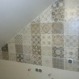 Ściana w łazience wyłożona dekoracyjnymi, kwadratowymi płytkami w odcieniach szarości i beżu, z widocznymi gniazdkami elektrycznymi i przygotowanymi otworami na instalację hydrauliczną.