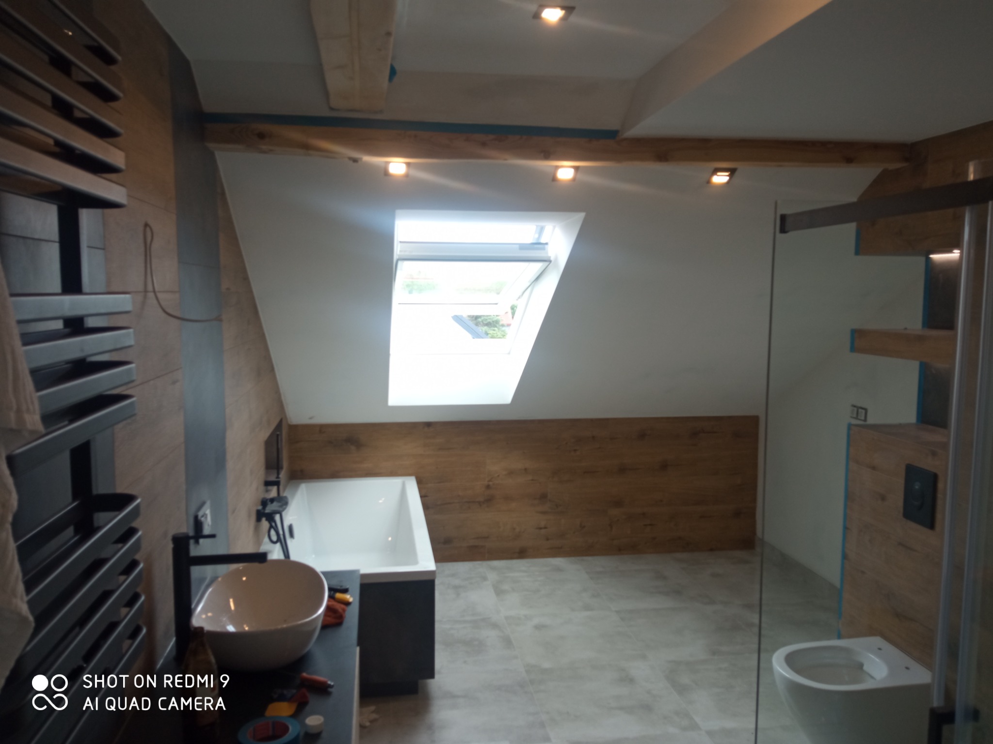 Nowoczesna łazienka na poddaszu z oknem dachowym, drewnianym wykończeniem ścian i szarą podłogą z płytek, widoczne narzędzia na blacie.