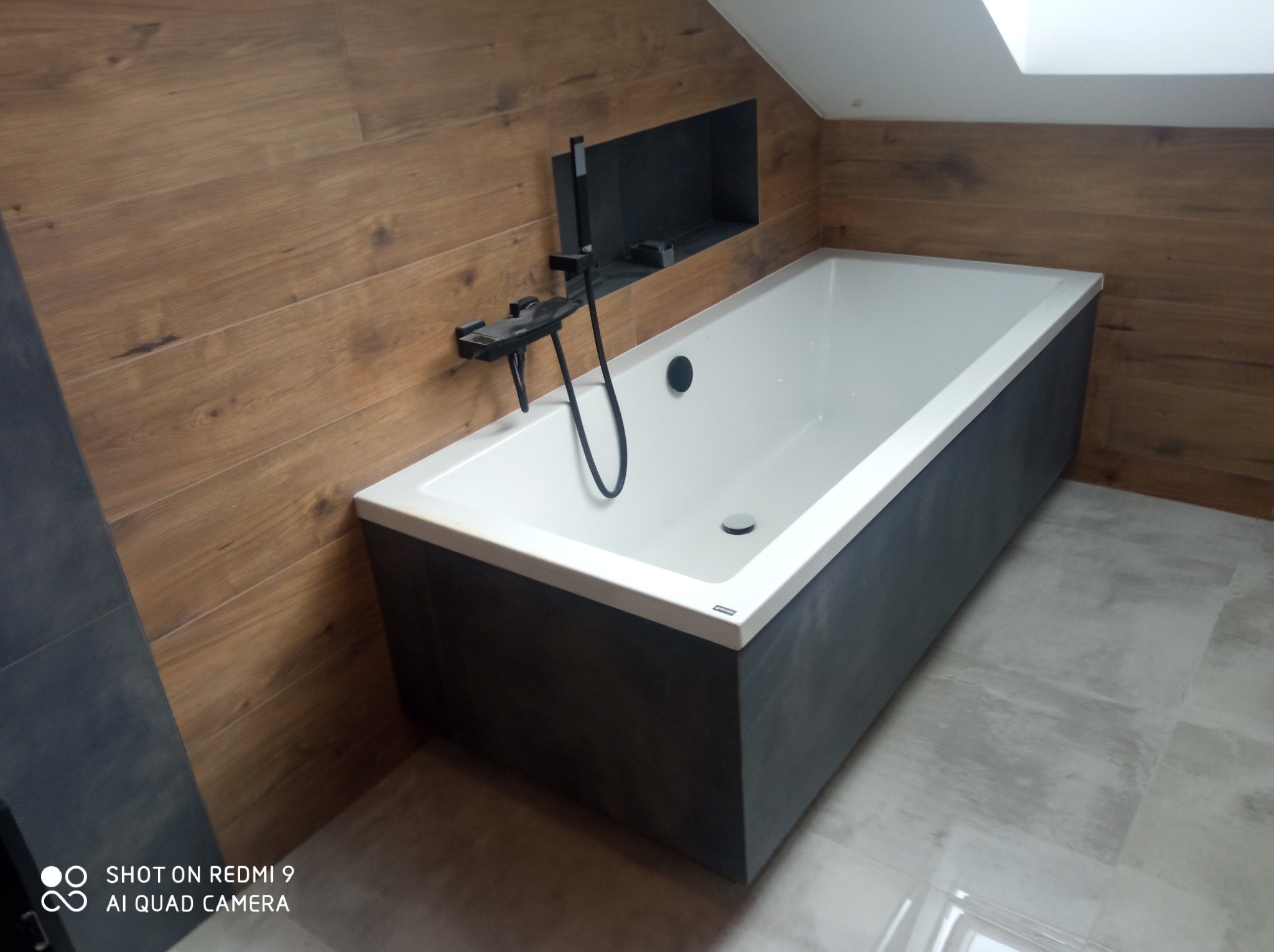 Nowoczesna wanna obudowana szarymi płytami, z czarną armaturą i wnęką w ścianie wyłożonej drewnopodobnymi płytkami, na podłodze szare płytki.