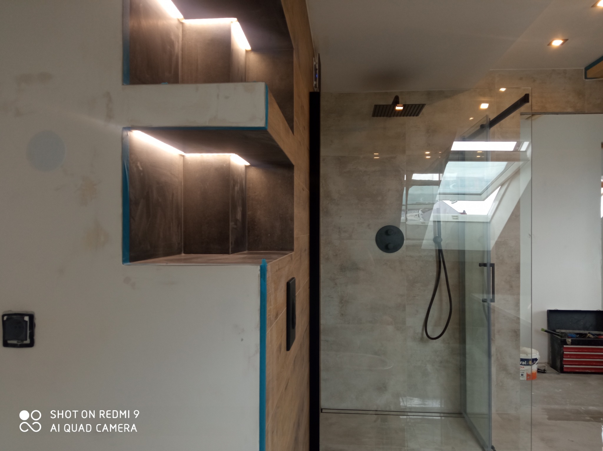 Fragment łazienki w trakcie prac wykończeniowych: zabudowa z podświetlanymi wnękami, przeszklona kabina prysznicowa z czarną armaturą i otwartym oknem dachowym, widoczne narzędzia i materiały.