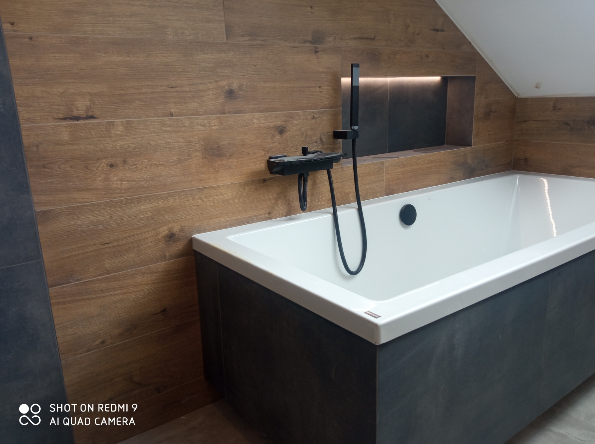 Nowoczesna łazienka na poddaszu z wanną obudowaną ciemnymi płytkami, drewnopodobne płytki na ścianach, czarna armatura i wnęka z podświetleniem LED.