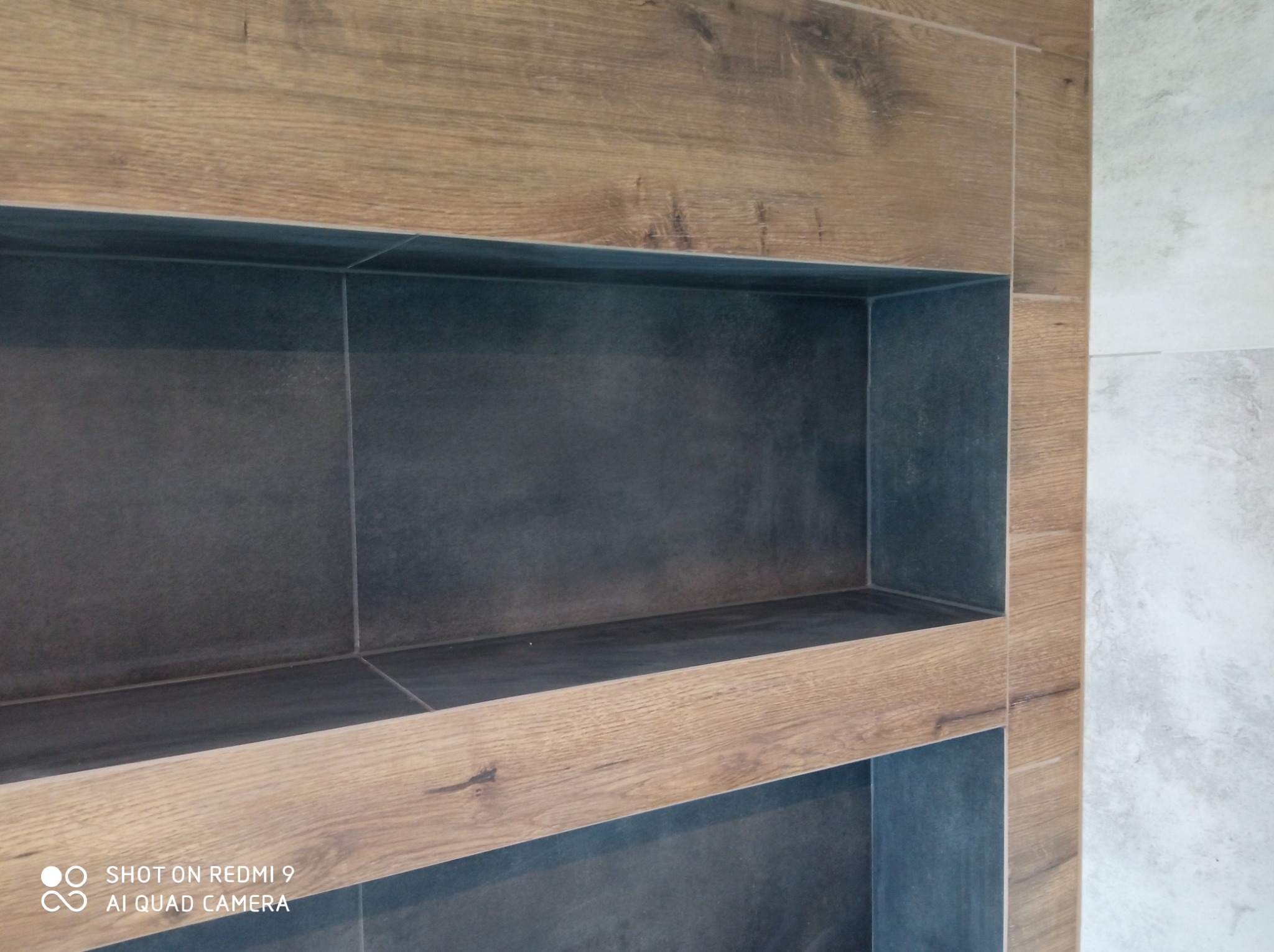 Narożna półka wnękowa wykończona płytkami imitującymi drewno, z ciemnoszarym wnętrzem, zamontowana na ścianie pokrytej jasnoszarymi płytkami.