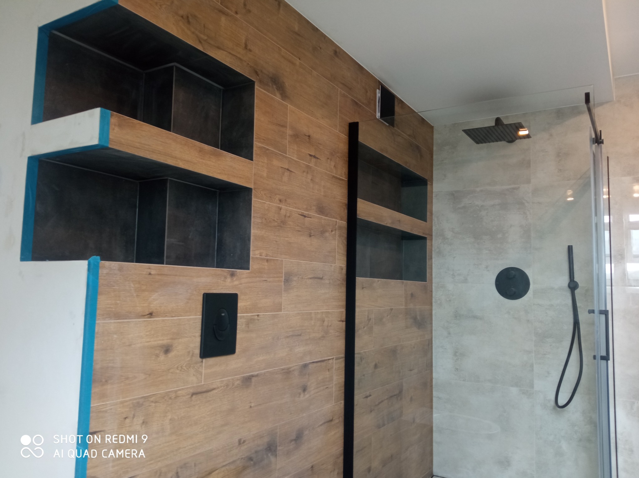 Nowoczesna łazienka z drewnopodobnymi płytkami, wnękami w ścianie wyłożonymi ciemną glazurą, czarną armaturą i szklaną kabiną prysznicową; fragment ściany w trakcie prac wykończeniowych...