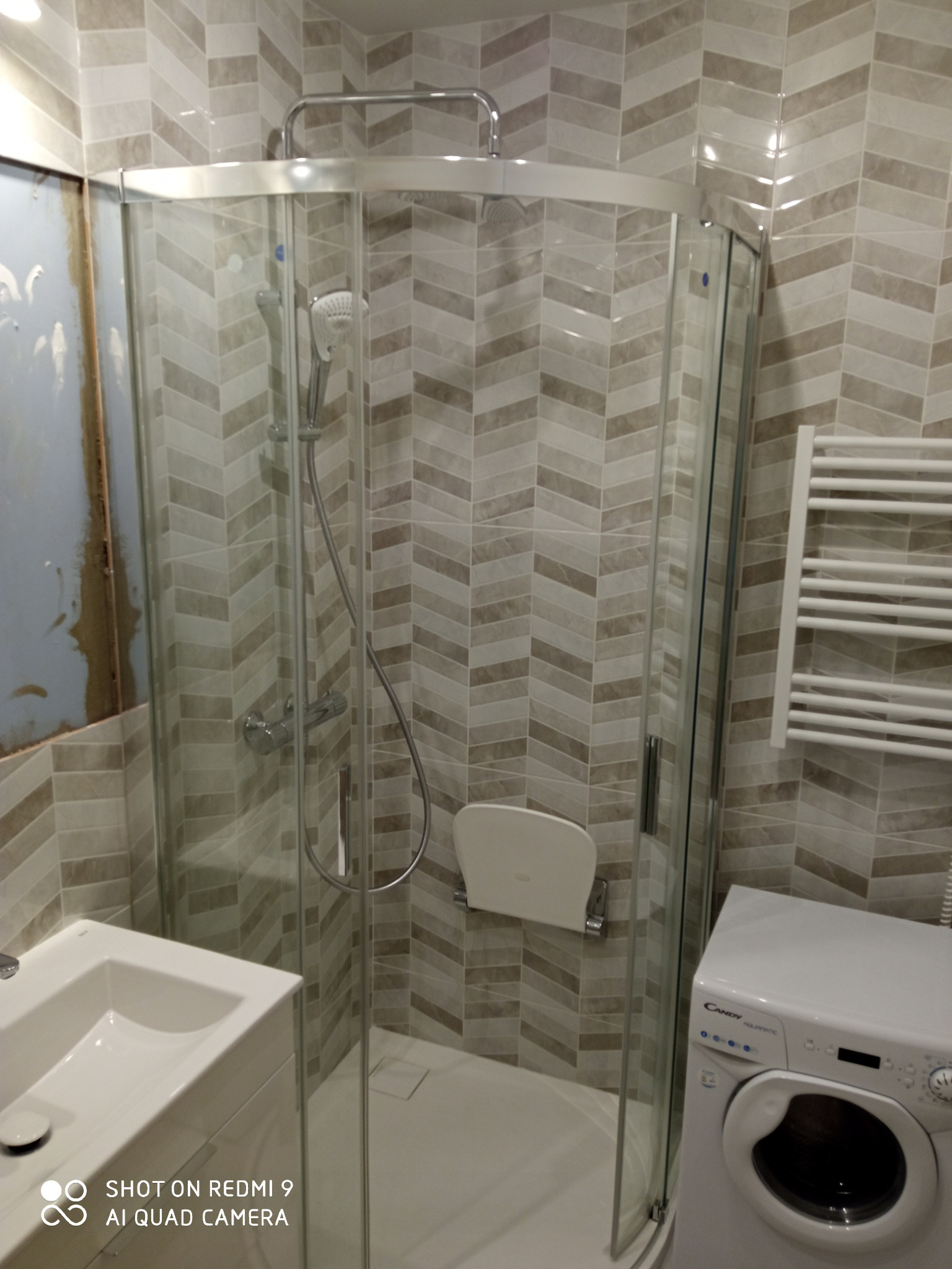 Kabina prysznicowa narożna z siedziskiem, grzejnik drabinkowy i pralka w łazience z chevronową glazurą; widoczny fragment ściany w trakcie prac wykończeniowych.