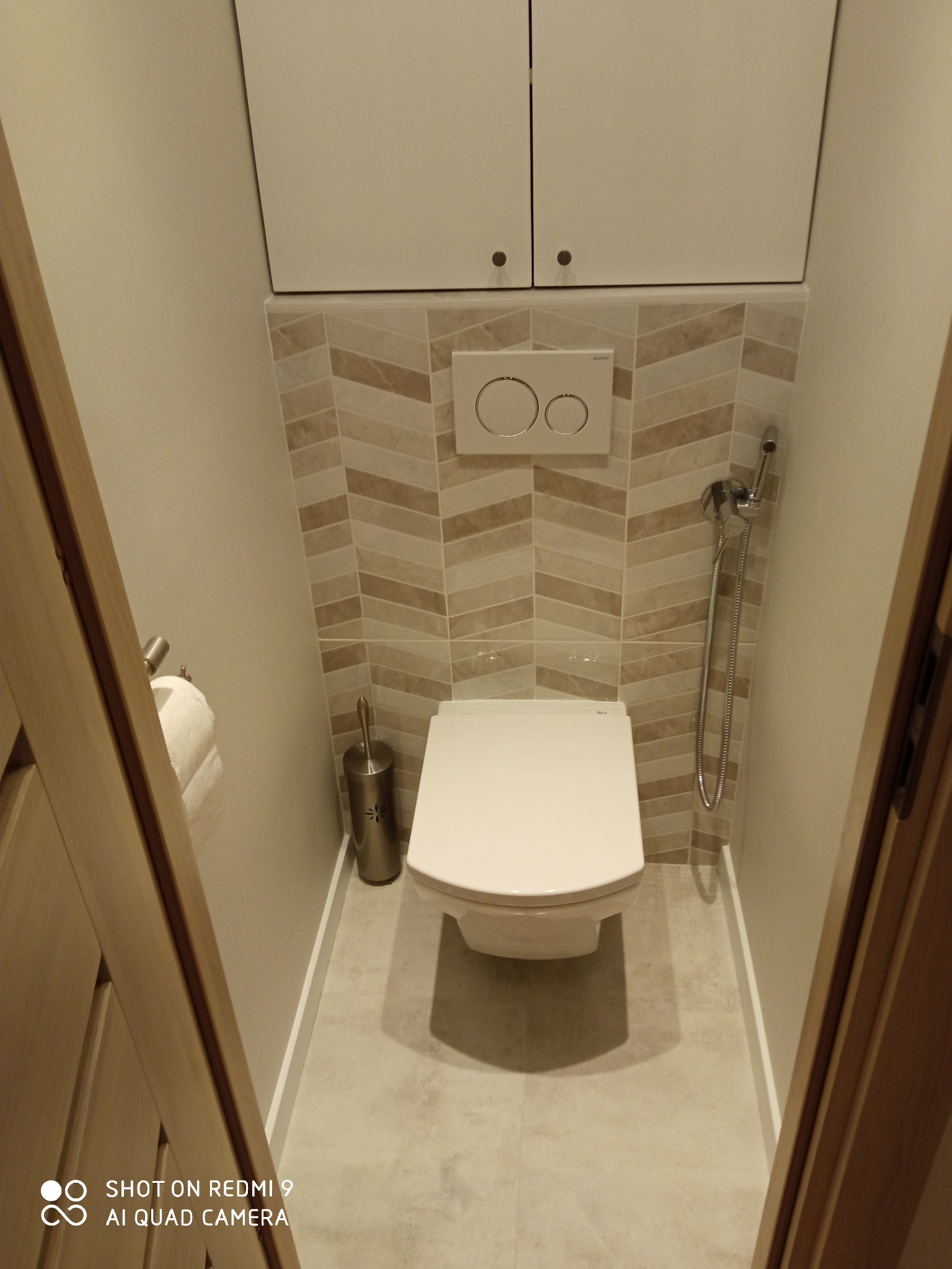 Toaleta w małej łazience w Rumi, z zabudową nad stelażem, wykończona płytkami w beżowe zygzaki, widoczny bidetta i szczotka do WC.