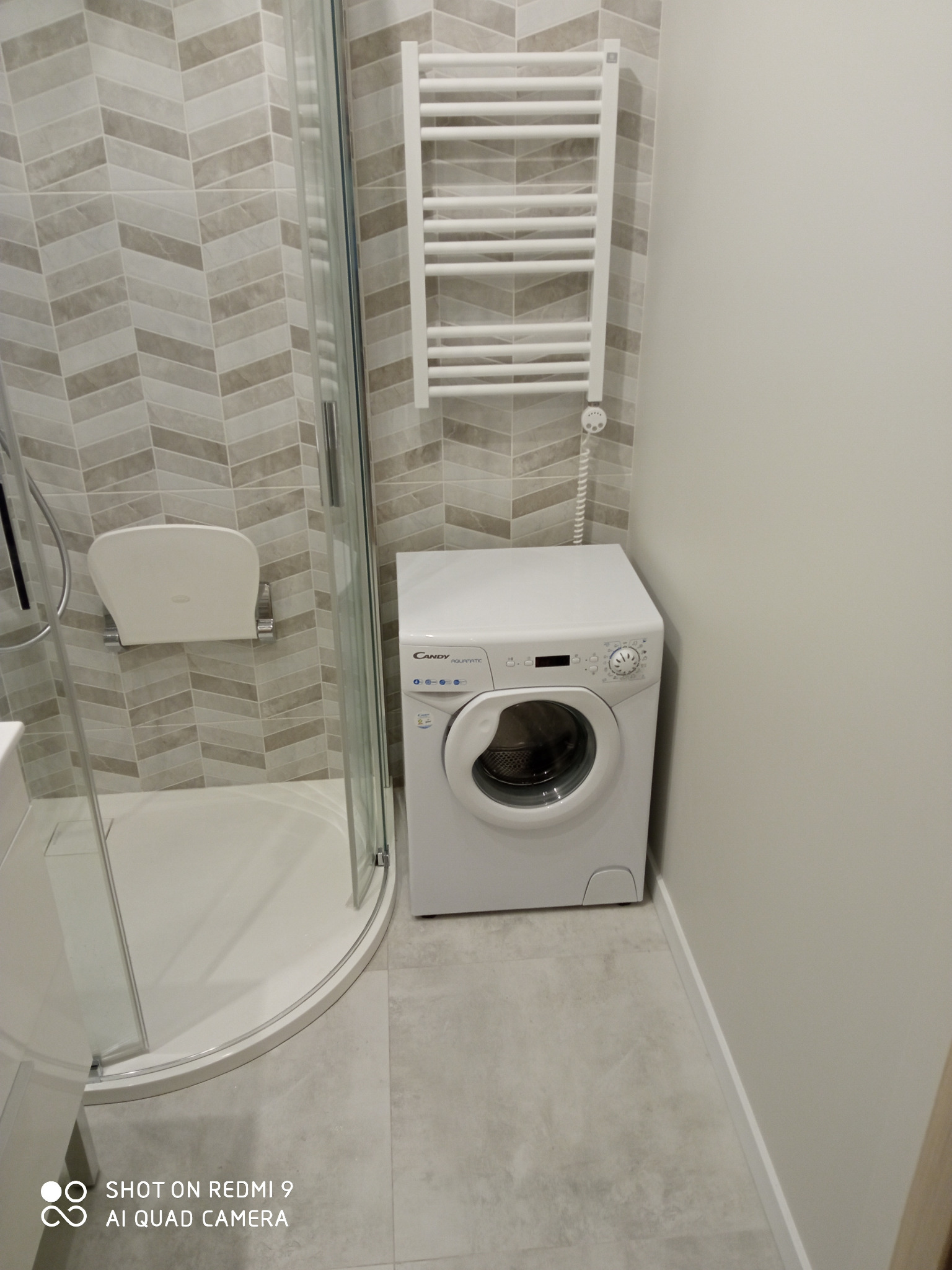 Nowoczesna łazienka z kabiną prysznicową z siedziskiem, grzejnikiem drabinkowym i pralką Candy Aquamatic na tle ściany wyłożonej płytkami w zygzaki.