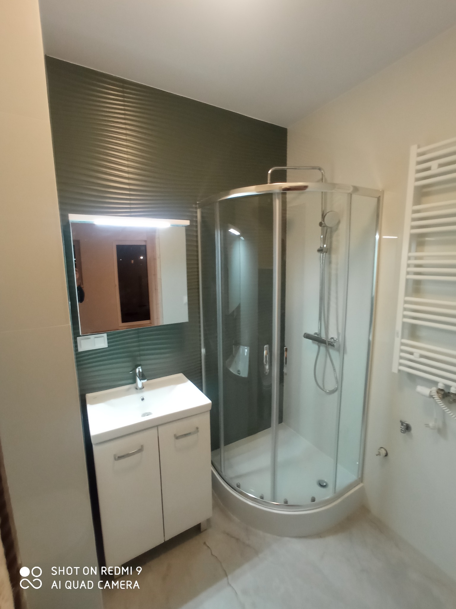 Wykończona łazienka z kabiną prysznicową narożną, białą szafką pod umywalką z lustrem podświetlanym LED i grzejnikiem drabinkowym na ścianie w kolorze złamanej bieli, z akcentem zielonej ściany...