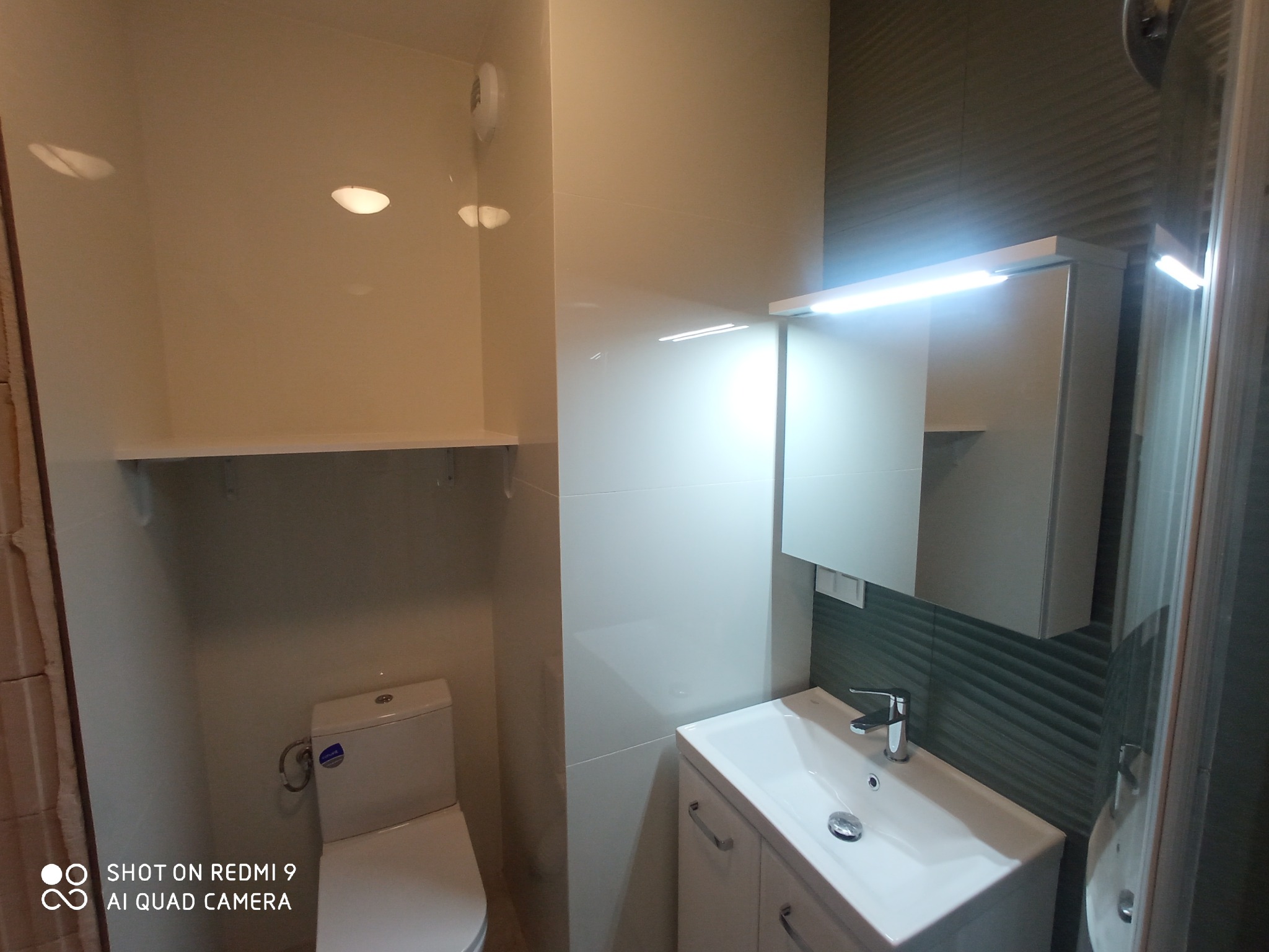 Jasna łazienka z toaletą, umywalką z szafką i lustrem z oświetleniem LED, wykończona białymi płytkami i dekoracyjnymi panelami ściennymi w odcieniach szarości.