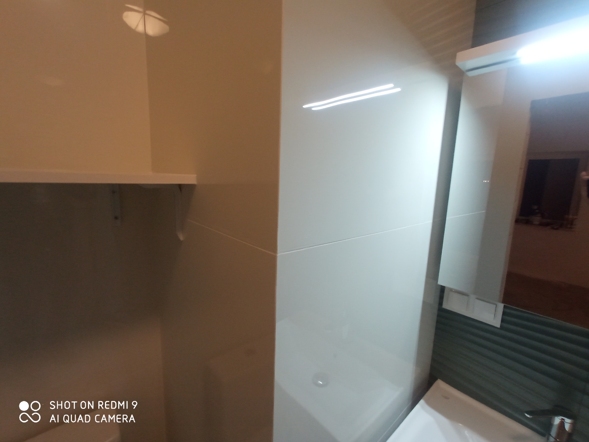 Fragment łazienki po remoncie: białe płytki na ścianach, umywalka w rogu, szafka wisząca nad umywalką, lustro z oświetleniem.