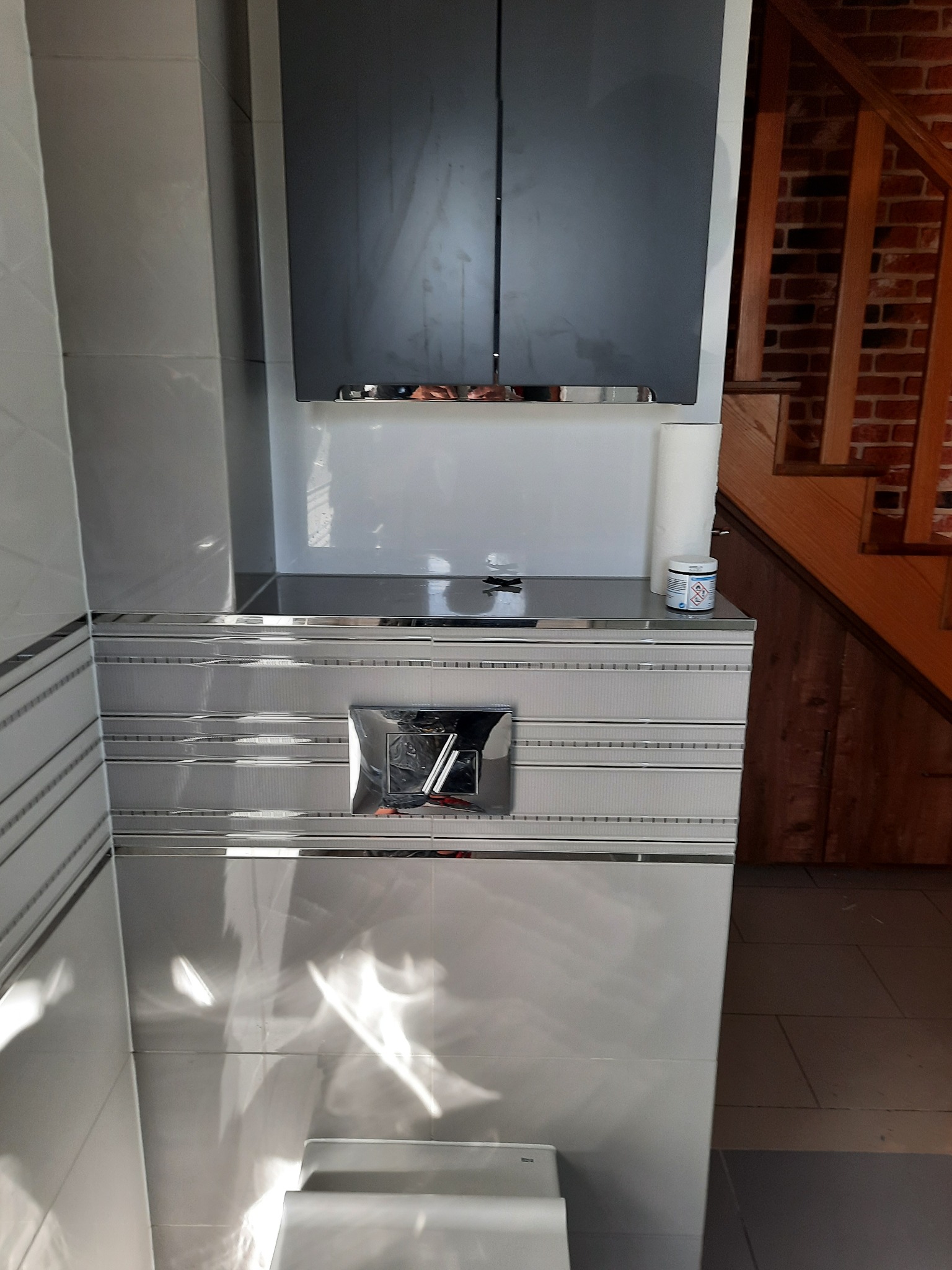 Szara szafka wisząca nad zabudowaną toaletą z chromowanym przyciskiem spłukującym, białe płytki ze srebrnymi paskami dekoracyjnymi, rolka papieru toaletowego i pojemnik na blacie, w tle fragment...