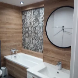 Nowoczesna łazienka z drewnopodobnymi płytkami na ścianach, ozdobną mozaiką, okrągłym lustrem i białą umywalką z szafką, oświetlona punktowymi lampami.