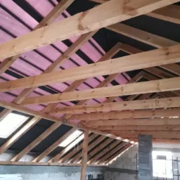 Konstrukcja więźby dachowej z widoczną membraną dachową w kolorze różowym, okna dachowe w trakcie montażu, surowy stan poddasza z bloczków betonowych i elementami szalunków.