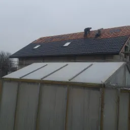 Praca na dachu: Dwóch dekarzy układa dachówkę na nowym dachu z oknami dachowymi, widok z dołu na tle szklarni i pochmurnego nieba.