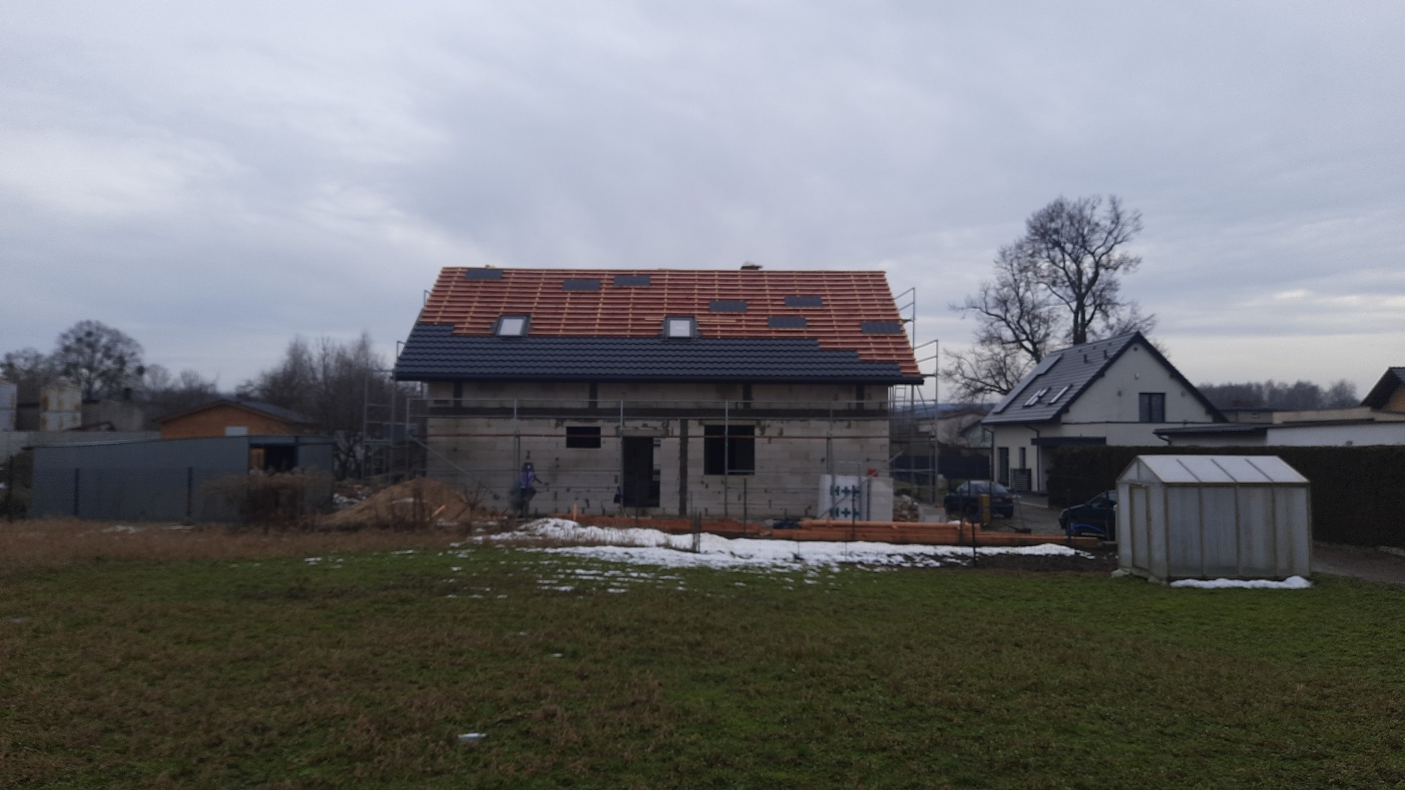 Budowa domu jednorodzinnego w trakcie: częściowo pokryty dachówką, widoczne rusztowania i przygotowane drewno, na pierwszym planie trawnik z resztkami śniegu.